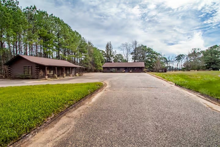 A wide asphalt driveway leads to two brown wooden buildings surrounded by pine trees and grassy areas under a partly cloudy sky.