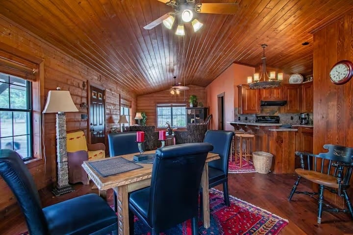 Cozy cabin interior with wood-paneled walls and ceiling, dining table with black chairs, a kitchen area, and warm lighting. A red rug and large windows complete the space.