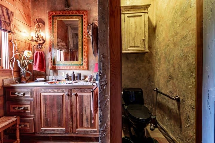 Rustic bathroom with wooden cabinetry, a large framed mirror, and antler wall sconce on the left; separate toilet area with a wooden cabinet on the right.