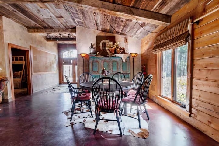 Rustic dining room with a round table and six chairs, wood-paneled walls, large window, animal hide rug, and vintage hutch against the far wall.