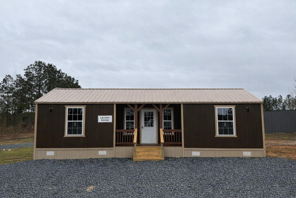 A small brown building with a metal roof, front porch, and a “Laundry Room” sign on the left window, surrounded by gravel and grass under a cloudy sky.